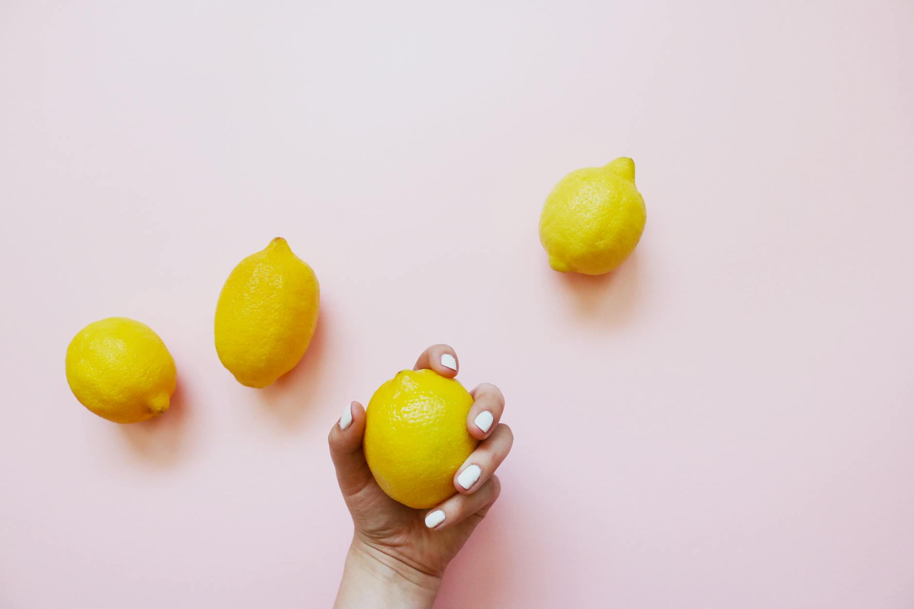 A hand holding a fresh lemon on a soft pink background surrounded by other lemons